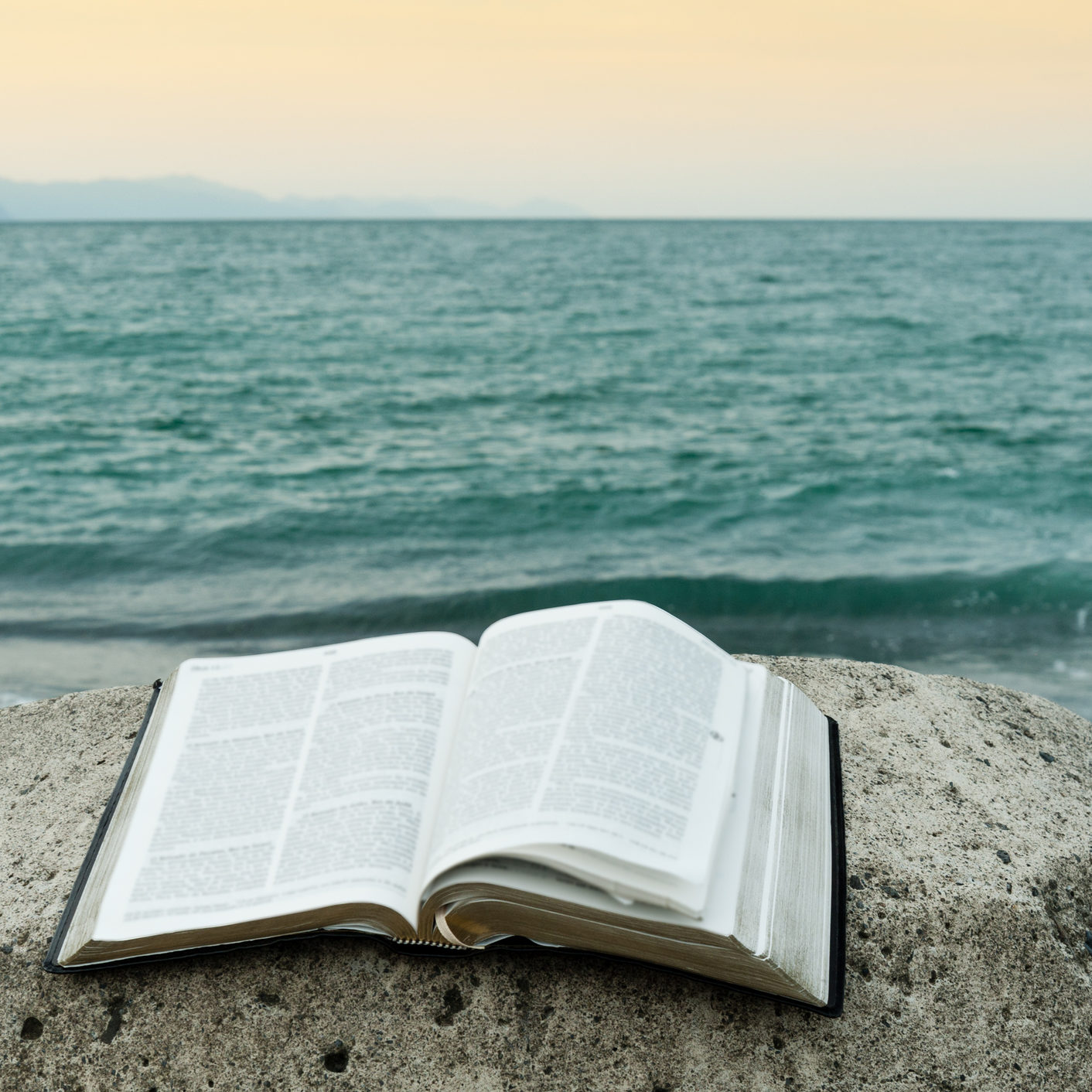 Open bible on top of a rock in front of the sea of greenish waters and an orange sky. Copy the space.